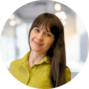 A woman with long brown hair is smiling, wearing a bright green blouse, while in the softly blurred background, lights hint at a modern indoor setting ideal for showcasing CRM implementation.