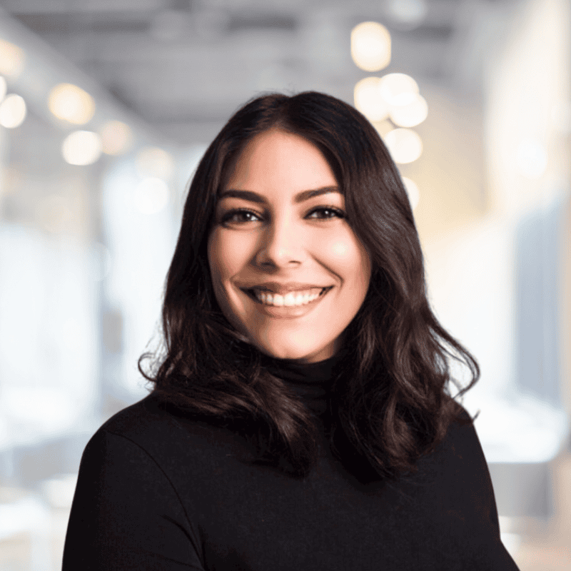 A person with long dark hair in a black turtleneck smiles warmly in a modern, softly lit office setting, representing Solutions Metrix—a leader in CRM consulting.