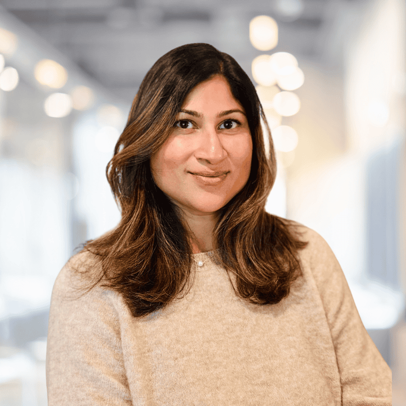 A person with shoulder-length hair and a soft smile poses in a warmly lit office environment at Solutions Metrix. The blurred background highlights modern lighting and glass elements, creating a professional yet relaxed atmosphere ideal for CRM implementation consulting.