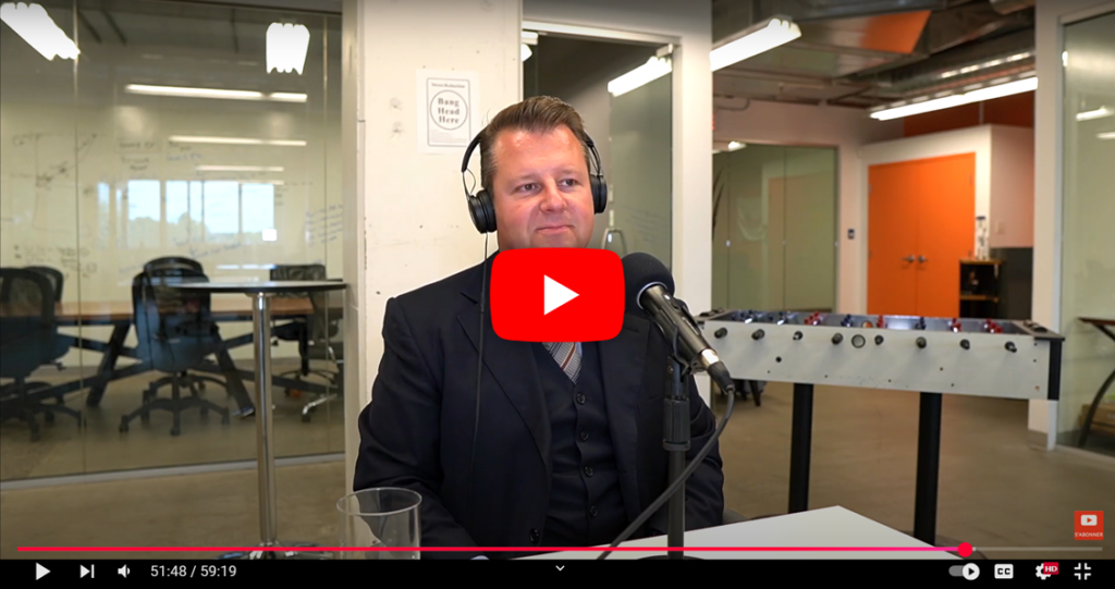 A man wearing headphones and a suit speaks into a microphone during a podcast recording in a modern office setting. A foosball table and glass walls with whiteboard scribbles are visible in the background. The video is paused at 51:48 in a YouTube player.